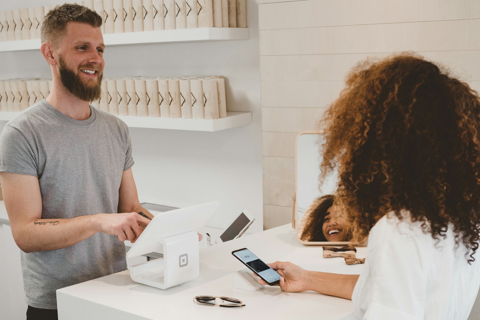 A shop owner at his counter smiles as a customer pays by phone.